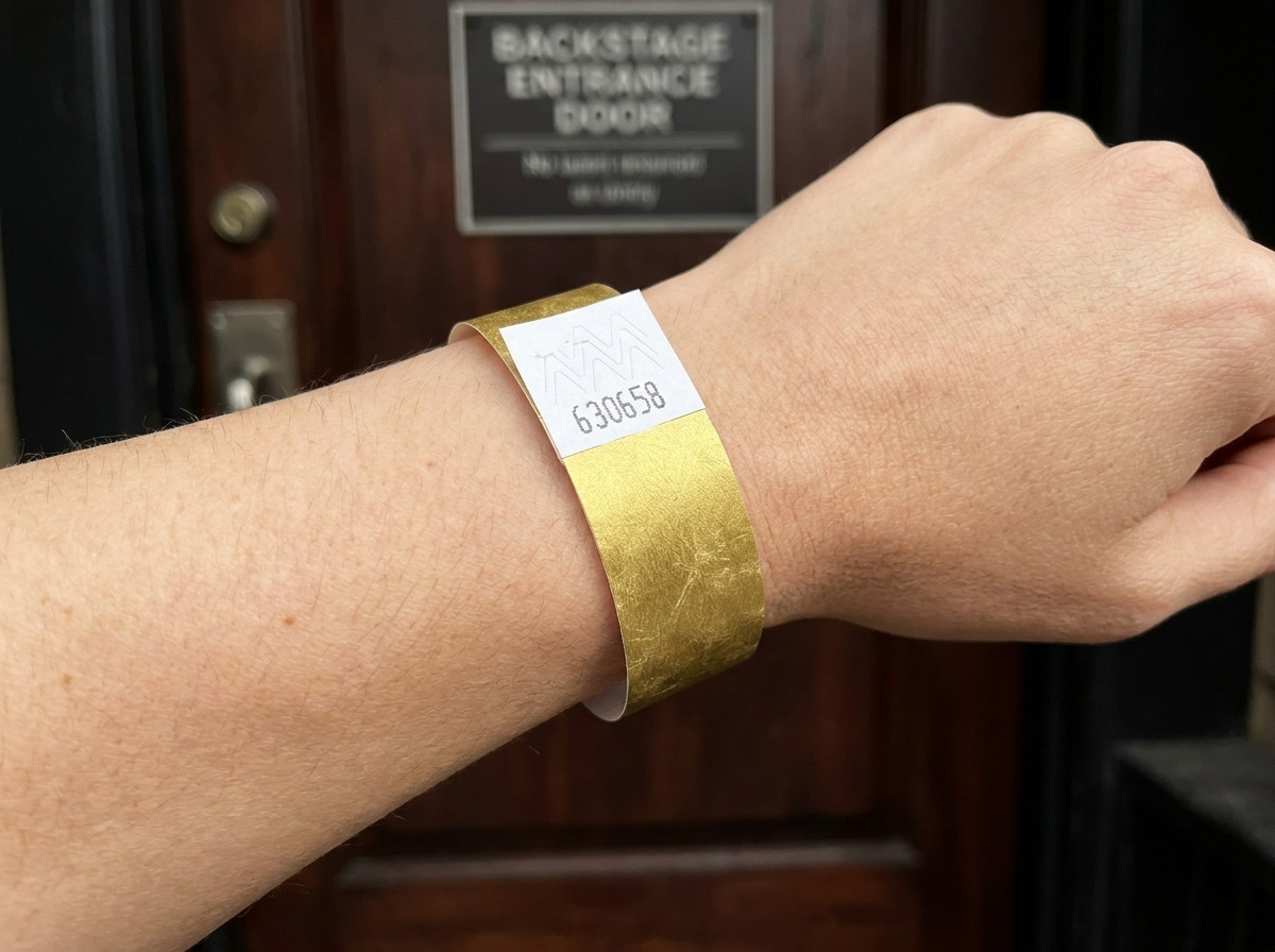 Bracelet VIP festival avec impression métallisée dorée et code-barres, porté sur un poignet devant une entrée backstage